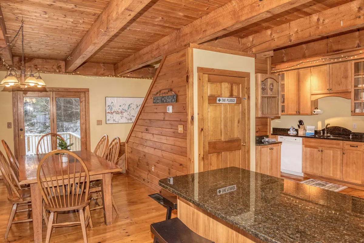 A warm, open-concept dining and kitchen area with a wooden dining table, kitchen island, exposed wooden beams, and expansive windows. The layout encourages togetherness, cooking, and shared meals, while the windows showcase the snowy scenery outside.