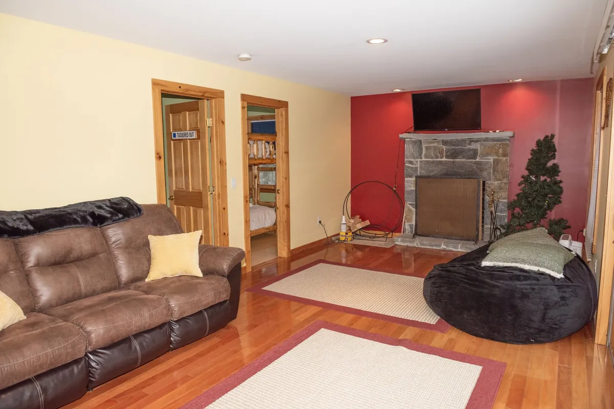 A cozy living room featuring a stone fireplace, a plush brown couch, a bean bag chair, and a bold red accent wall. The arrangement invites families to relax, play games, or watch TV together by a glowing fire.