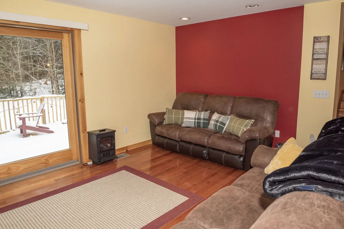 A cozy living space featuring comfortable sofas arranged near a large window and glass door that leads out onto a deck. The view outside hints at greenery and the inviting outdoors. The interior exudes warmth, while the window visually connects the inside space to the natural setting beyond.