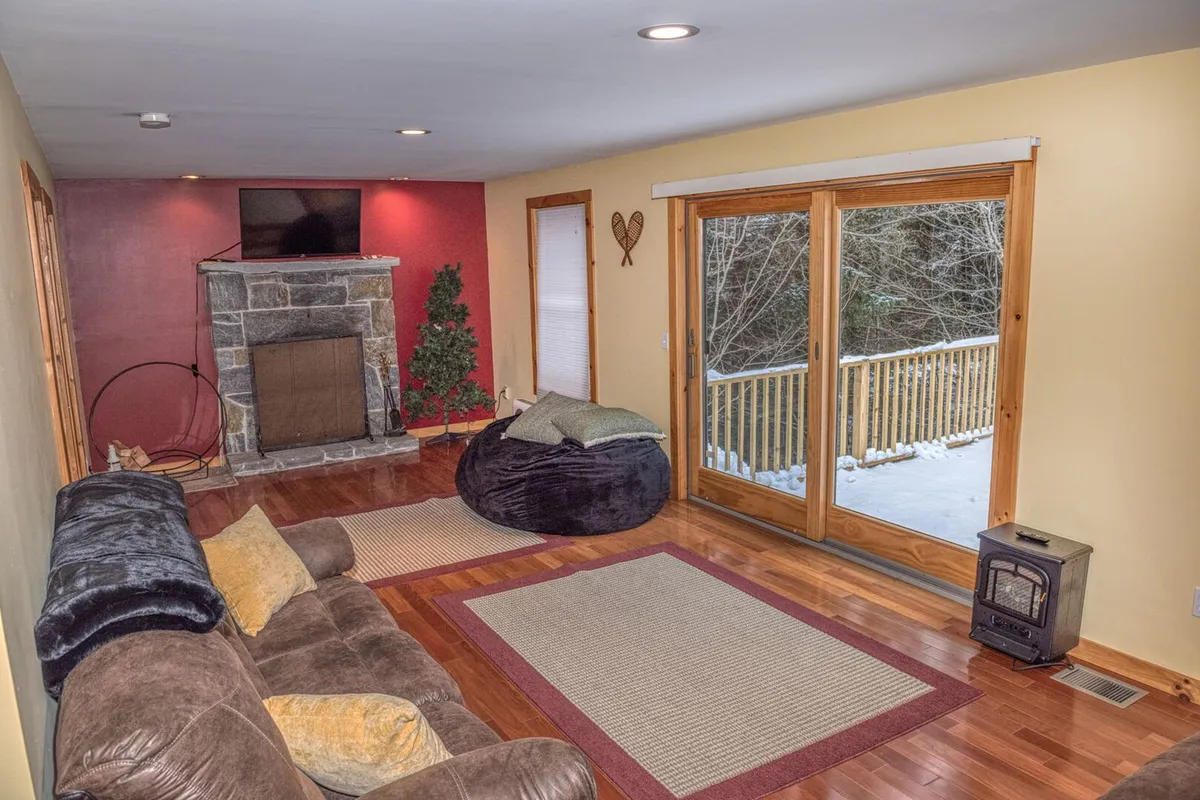 A spacious, sunlit living room featuring a striking stone fireplace, plush sofa, bean bag chair, and large windows that frame beautiful views of the snowy forest outside. The inviting setup is ideal for relaxing, gathering, and enjoying the ambiance of the fire.