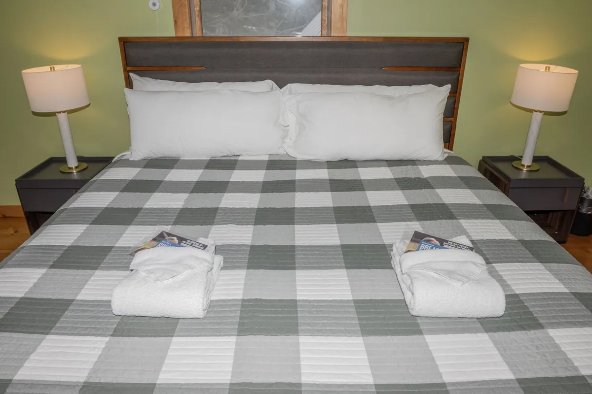 A cozy bedroom featuring a comfortable bed with plush bedding, flanked by nightstands and lamps. Towels and magazines are neatly arranged, and natural light streams in through the window, creating a tranquil, restful ambiance.