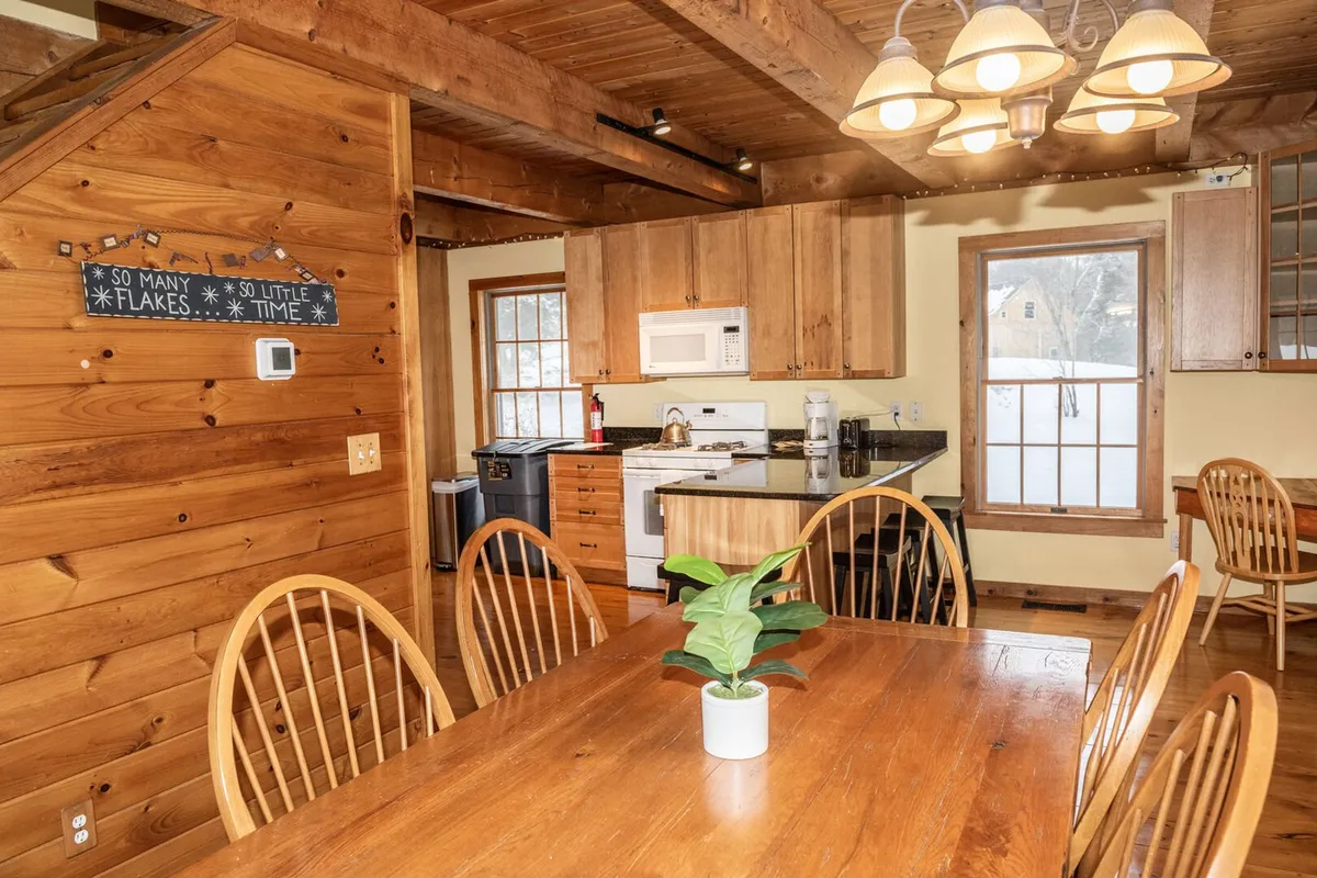 A warm, inviting dining area with a large wooden table, exposed beams, and kitchen cabinetry in the background. The table is centrally arranged for group gatherings, with a decorative sign adding a touch of local character. The lighting is soft, giving the space a festive and communal atmosphere.