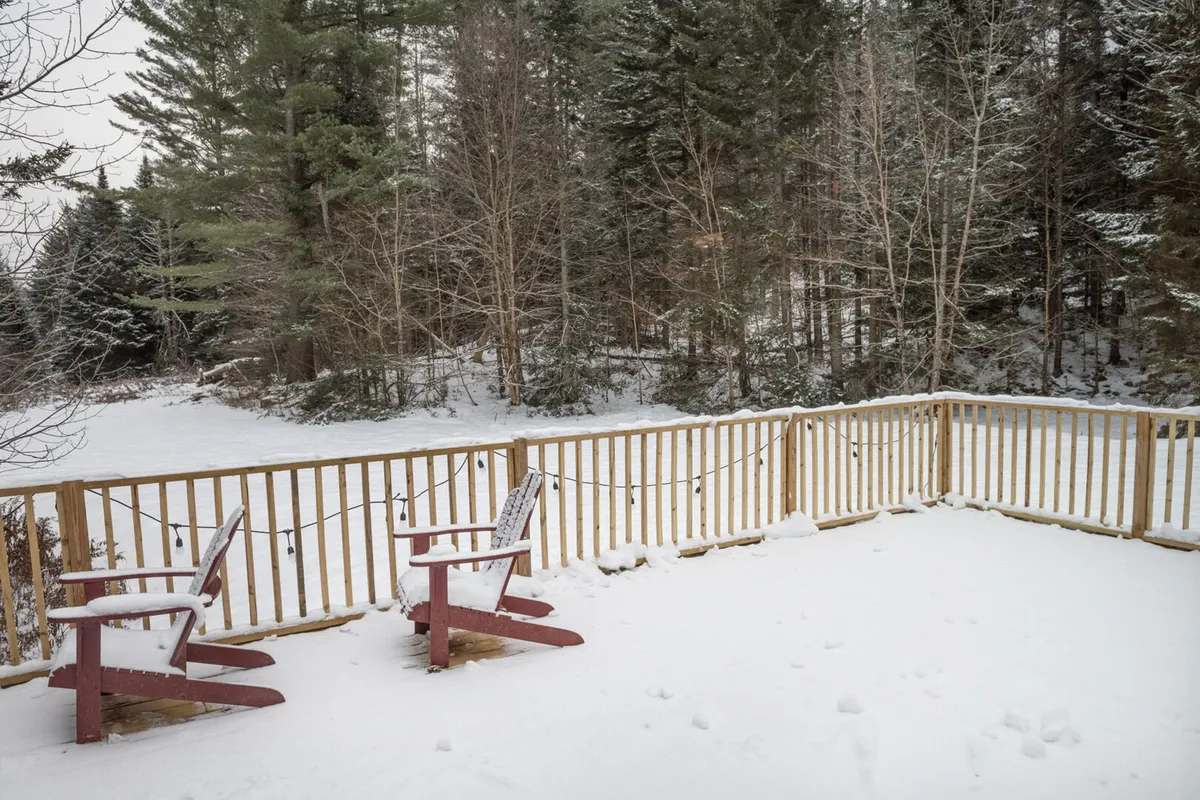 A tranquil snow-covered deck with two bright red Adirondack chairs facing out towards a forest of trees. The scene is quiet and inviting, ideal for relaxing, sipping hot drinks, or watching the snowfall. The untouched wilderness immediately beyond the deck highlights the home’s immersion in nature.