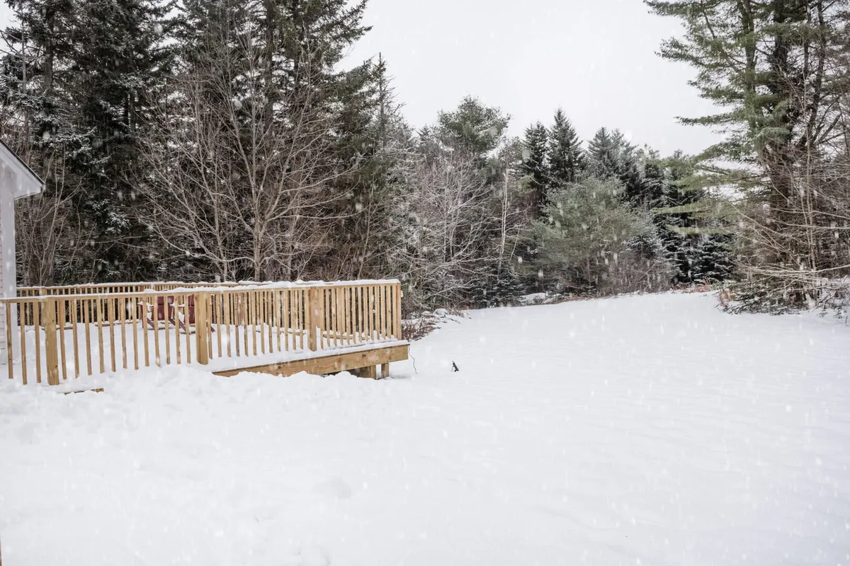 A serene winter landscape featuring a wooden deck looking out onto snow-covered ground, surrounded by tall, leafless trees. The deck is partially dusted with snow, suggesting recent snowfall and inviting coziness within the scene. The natural wooded surroundings evoke privacy, seclusion, and the beauty of Vermont’s outdoors right at your door.