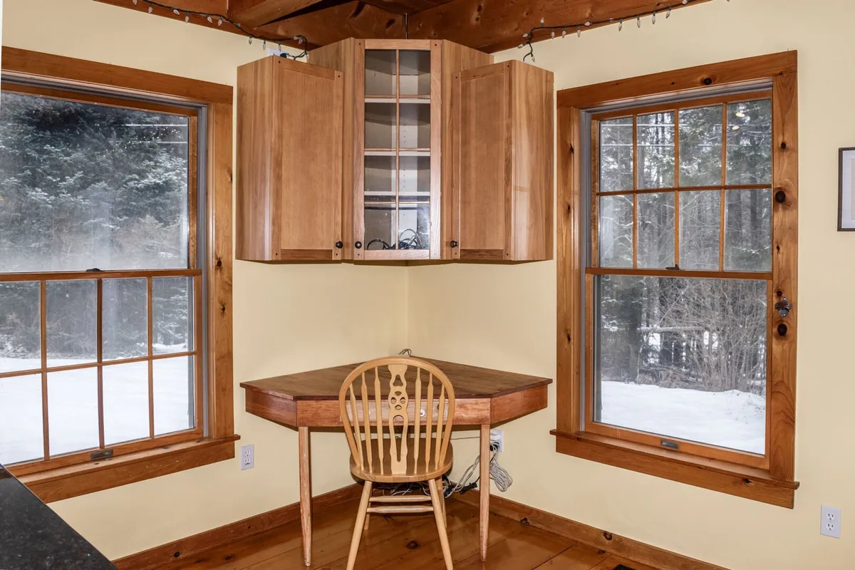 A wooden desk is positioned beside a window, offering a tranquil view of the surrounding forest. Sunlight streams in, illuminating the workspace and providing a calming natural backdrop. The setting is quiet and free of distractions, ideal for focused work or reading.