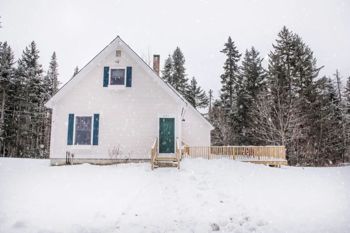 An inviting exterior shot of the white house with a green door and a spacious deck area, all surrounded by snowy landscape and trees. The view shows the home’s proximity to vast open areas and hints at the start of trails leading into the woods.